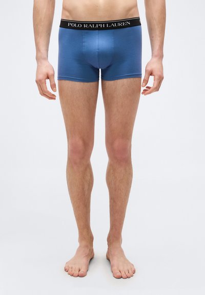 Άνδρας όρθιος, ξυπόλητος, φοράει μπλε boxer briefs Polo Ralph Lauren με μαύρη μέση, πάνω σε απλό λευκό φόντο.