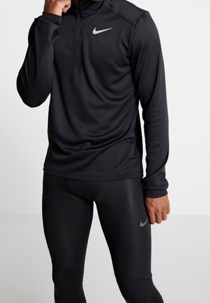 Mężczyzna noszący czarną bluzę Nike z długim rękawem na zamek oraz czarne sportowe legginsy Nike, stoi na jednolitym białym tle.