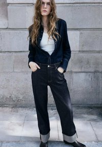 Giacca di denim scuro con bottoni, canottiera bianca, jeans scuri larghi con orli risvoltati, indossati con stivaletti marroni, in piedi contro un muro di pietra.