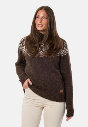 Donna con lunghi capelli castani che indossa un maglione marrone a fantasia lavorato a maglia e pantaloni beige chiaro, una mano in tasca, che guarda leggermente di lato.