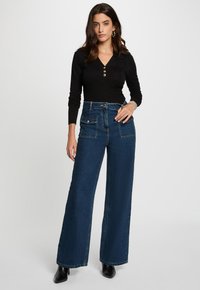 Jeans a gamba larga blu scuro a vita alta con chiusura a bottone, passanti per la cintura e tasche frontali. Indossati con un top nero a maniche lunghe.