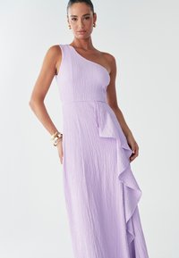 En axelklänning i texturerat ljuslila tyg, med en veckad detalj längs sidan och en figurnära midja. Mjuk, flödande siluett.