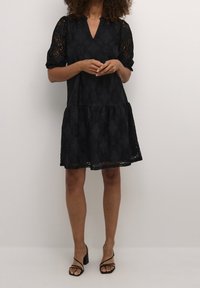 Svart spetsklänning med V-ringning, korta puffärmar och en lager-på-lager kjol. Har intrikata blommönster och en genomskinlig textur. Svarta remsandaler.