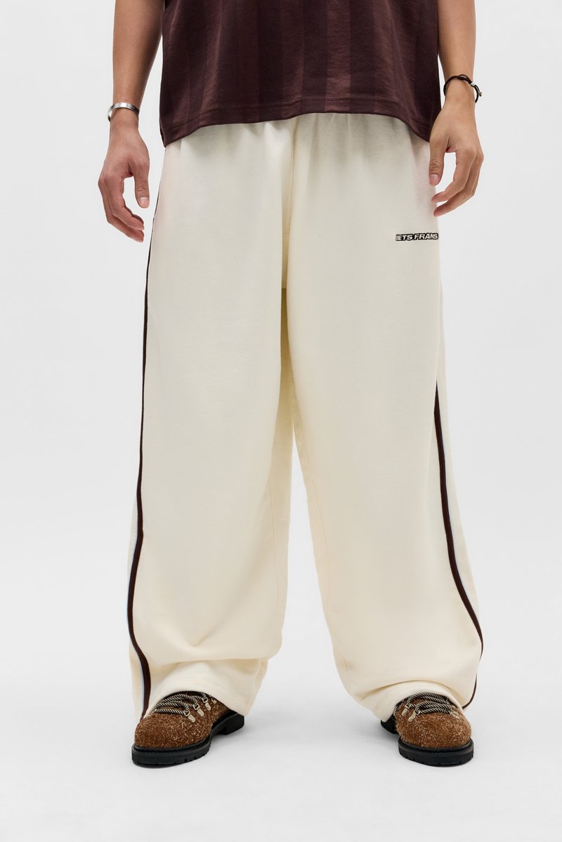 Άτομο που φοράει άνετο λευκό παντελόνι με μαύρες ρίγες στα πλάγια και καφέ παπούτσια με υφή, στέκεται με τα χέρια χαλαρά δίπλα στο σώμα.