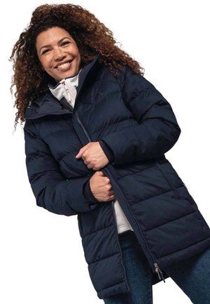 Navy blue puffer coat with a hood, zip closure, and large quilted sections. Paired with a light-colored inner layer and denim.