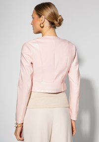Giacca in pelle rosa cropped con maniche lunghe, caratterizzata da dettagli sulle cuciture posteriori e una finitura lucida. Indossata sopra un top beige.