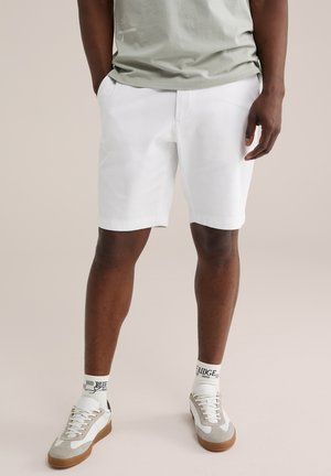 Mężczyzna ubrany w białe spodenki, jasnoszarą koszulkę, białe skarpetki z nadrukiem oraz białe i beżowe trampki, stojący z rękami w kieszeniach.