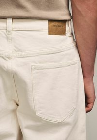 Hvide denimbukser med en tekstureret overflade, udstyret med et brunt lædermærke bagpå med "DENIM SUPPL. Co" præget.