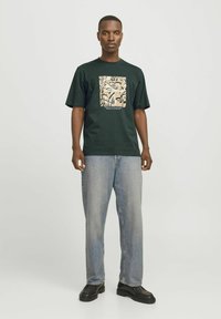 Camiseta de algodón verde oscuro con un estampado gráfico cuadrado en beige con diseños abstractos, combinada con jeans de mezclilla azul claro y zapatos negros.