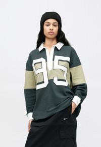 Πράσινη και μπεζ μακρυμάνικη πόλο μπλούζα με λευκές λεπτομέρειες και μεγάλο '95' στο μπροστινό μέρος. Φορεμένη με μαύρο σκουφί και φαρδιά μαύρα παντελόνια.