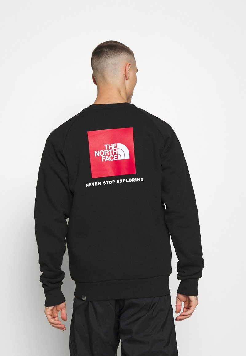 Homme aux cheveux courts portant un sweat-shirt noir avec un logo carré rouge et le texte blanc "THE NORTH FACE" et "NEVER STOP EXPLORING" au dos.