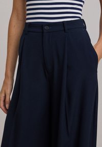 Granatowe spodnie szerokimi nogawkami o dopasowanym kroju, gładkiej fakturze, subtelnych plisach i guzikach w talii; kieszenie po obu stronach.