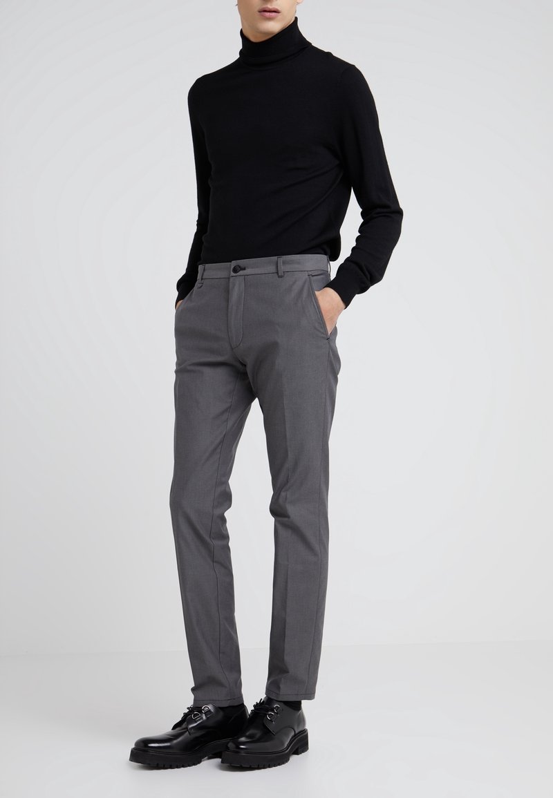 Homme portant un pull à col roulé noir, un pantalon gris sur mesure et des chaussures en cuir noires, debout les mains dans les poches sur fond blanc.