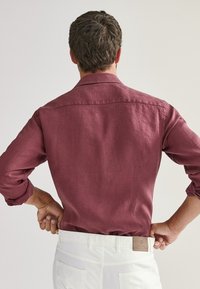 Burgundowa lniana koszula z kołnierzykiem i długimi rękawami, o luźnym kroju. Noszona z białymi spodniami, które podkreślają teksturowany materiał.