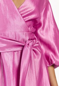 Roze satijnen jurk met een V-hals, brede pofmouwen en een strikdetail in de taille. De stof heeft een gladde, glanzende textuur.