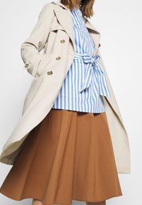 Béžový trenčkot se zlatými knoflíky, modro-bíle pruhovaná košile s detailem kravaty a hnědá rozšířená sukně po kolena. Jemné textury.