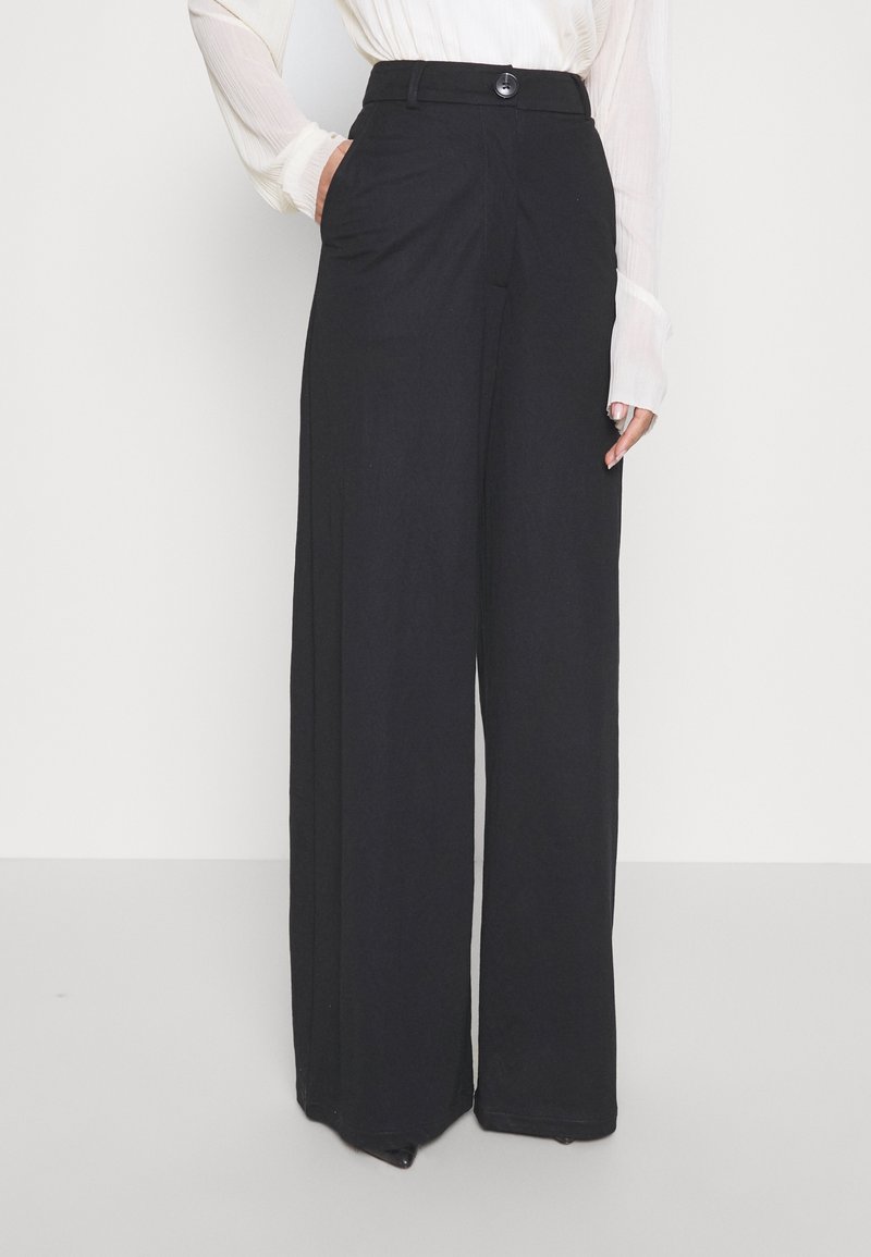 Femme portant un pantalon noir taille haute à jambes larges avec poches avant et un chemisier blanc à manches longues, debout devant un fond uni.