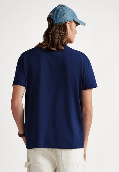 Μπλούζα από βαμβάκι σε ναυτικό μπλε χρώμα με κοντά μανίκια, στρογγυλή λαιμόκοψη, λεία υφή. Φορεμένη με ανοιχτό μπλε τζιν καπέλο, παρουσιάζοντας την πίσω όψη.