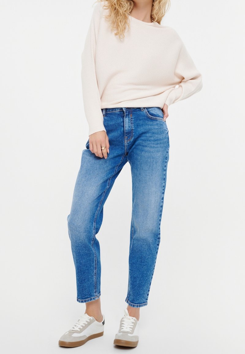 Vaaleanpunainen ribbineulottu neulepaita, siniset korkeavyötäröiset farkut ja valkoiset tennarit ruskeankeltaisilla yksityiskohdilla. Neulepaidassa on leveä pääntie; farkut ovat suoraa lahjetta.