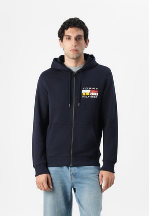 Hoodie zippé marine en tissu doux avec poches avant et un design de logo en rouge, jaune, bleu et blanc sur la poitrine.