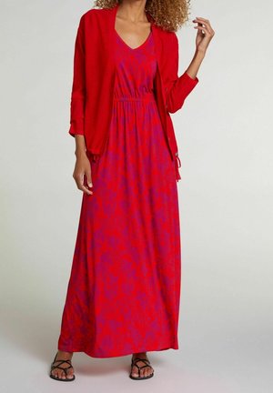 Kobieta w czerwono-fioletowej wzorzystej sukience maxi, czerwonym kardiganie i czarnych sandałach na jasno jednolitym tle.
