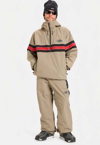 Khaki tuulekindel jope punaste ja mustade horisontaalsete triipudega, sobitatud samast materjalist pükstega. Materjal tundub kerge ja tekstuuriga. Jalga on pandud mustad saabased.