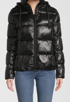 Kobieta nosząca błyszczącą czarną kurtkę puchową z kapturem i zamkiem z przodu, w parze z niebieskimi dżinsami na jednolitym tle.