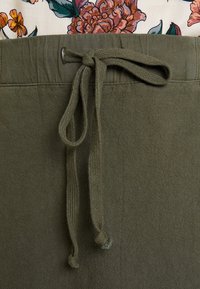 Tissu vert olive avec un tissage texturé, doté d'un lien de serrage à l'avant. La taille comporte une boucle cousue pour l'ajustement.