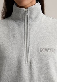 Helles graues Sweatshirt mit halbem Reißverschluss aus einem Baumwollmischgewebe. Verfügt über einen hohen Kragen und ein gesticktes "MO'PD"-Logo auf der Brust.