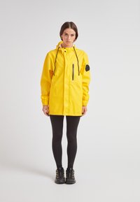 Keltainen vedenpitävä takki hupulla, jossa on musta vetoketjupussi ja kumilogo, päällä mustat legginsit ja vaelluskengät.