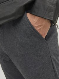 Grå, texturerade tygbyxor med en sidoficka, delvis täckta av en svart läderjacka, som visar en närbild av materialen.