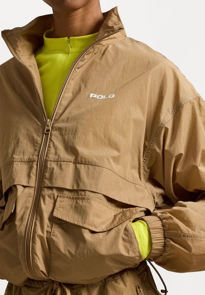 Gaiši brūna viegla jaka ar rāvējslēdzēju, striķi jostas vietā un kabatām; apakšā valkāta spilgti zaļa augsta kakla blūze.