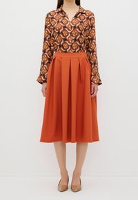 Mönstrad blus med orange och bruna virvlar, kombinerad med en enfärgad orange veckad midikjol. Spetsiga beiga klackar fulländar looken.