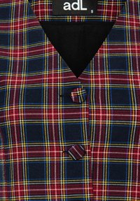 Rutig väst i marinblått, burgundy, gult och blått; har en V-ringning och en enkel knappstängning. Mjuk textil med en strukturerad yta.