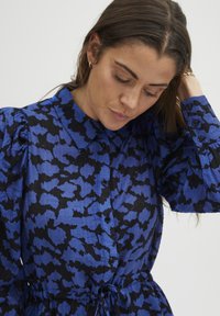 Blau und schwarz gemustertes Kleid mit Knopfleiste, Kragen und gerafften Ärmeln. Verfügt über eine verstellbare Taille mit Kordelzug.