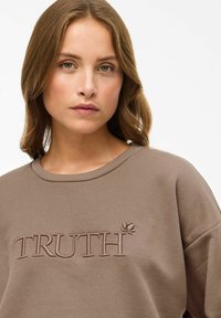 Brun sweatshirt med ribbat halslinning och broderad text "SANNING". Mjuk textur, avslappnad passform, med en bladaccent bredvid bokstäverna.