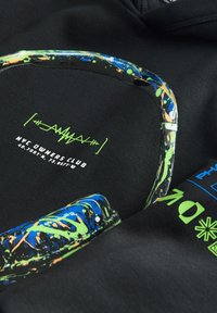 Fekete anyag neon zöld és kék festékfröccsenéses szemüveg grafikával és a „NYC Owners Club” felirattal, koordinátákkal alatta a ruházaton.