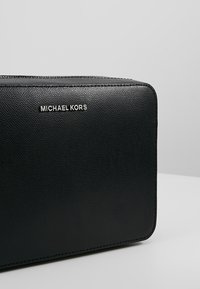 Borsa in pelle nera a forma rettangolare con texture di Michael Kors, con logo argentato e cerniera superiore, su uno sfondo grigio uniforme.