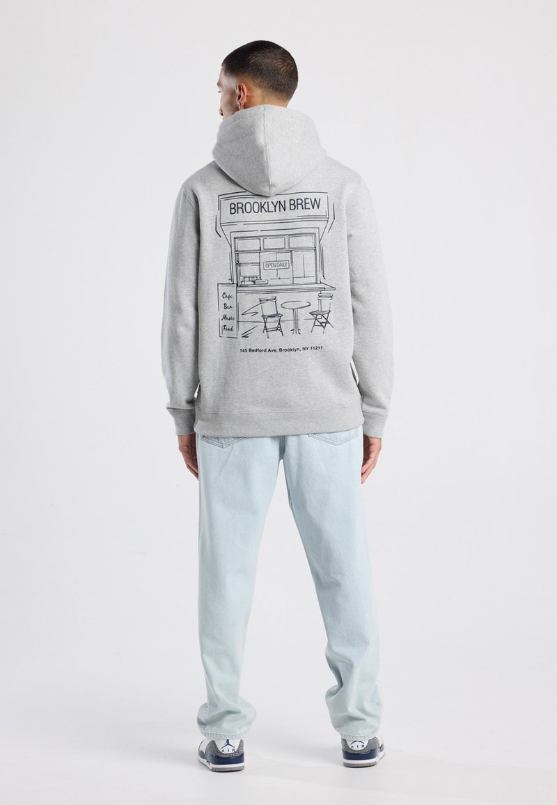 Felpa grigia con stampa grafica di un caffè sulla schiena, abbinata a jeans azzurri chiari e sneakers. Tessuto morbido e vestibilità casual.