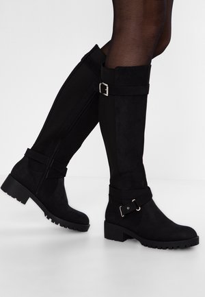 Bottes noires montantes avec talons épais et boucles, portées avec des collants noirs transparents, montrées sur des jambes croisées sur fond blanc.