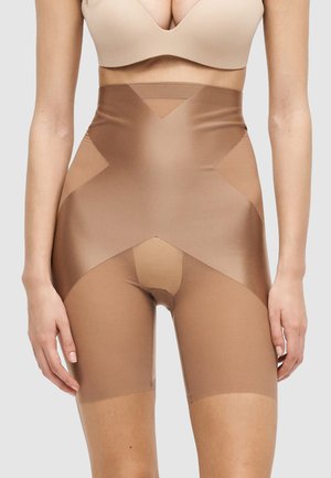 Žena v telových tvarovacích šortkách s vysokým pásom a béžovej podprsenke stojaca s uvoľnenými pažami pozdĺž tela na jednoduchom pozadí.