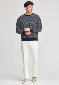 Fyrkantig sweatshirt i marinblå och vit randig design med ribbade ärmslut, ihop med krämfärgade byxor och beige skor, bärs av en stående manlig modell.