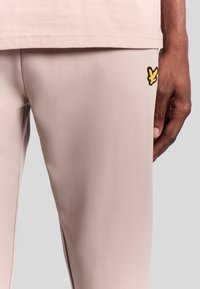 Beige atletiske bukser lavet af glat stof, med et lille sort og gult logo på låret. Nærbillede fremhæver tekstur og design.