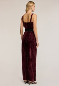 Burgundská sametová maxi šaty s tenkými ramínky a černým páskem, zdobené vertikálními pruhy v látce pro texturu a pohyb.