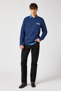 Young man wearing a blue Wrangler sweatshirt over a denim shirt, black jeans, and black boots, standing against a light background.