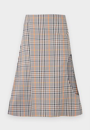 Nainen pukeutuneena oliivinvihreään yhden hihan pulleaan toppiin ja ruudulliseen hameeseen, jossa suuri reiden leikkaus, yhdistettynä mustiin remmisiin korkeakorkoisiin sandaaleihin.