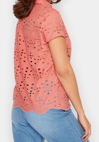 Kobieta w koralowo-różowej bluzce z krótkim rękawem z ażurowym wzorem oraz jasnoniebieskich dżinsach, pokazana z tyłu i lekko obrócona na bok.