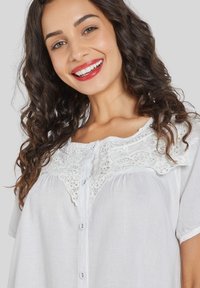 Blusa blanca con un delicado canesú de encaje, mangas cortas, cierre de botones en la parte delantera y una textura ligera. El encaje presenta patrones intrincados.