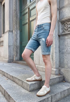 Personne portant un débardeur blanc, un short en jean et des baskets blanches, debout sur des marches en pierre près d'un bâtiment aux murs en pierre sculptée et à une porte verte.