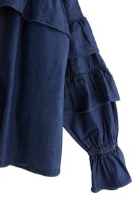 Mørkeblå denimbluse med flæser på ærmerne, samlede manchetter og en glat tekstur. Casual, afslappet pasform.